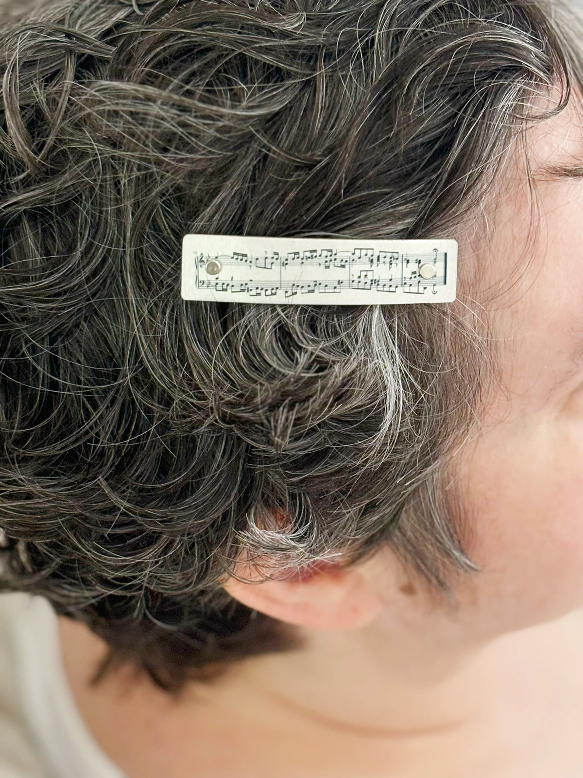 A close-up of a person's short, curly gray hair with a small rectangular silver hair clip featuring printed musical notation.