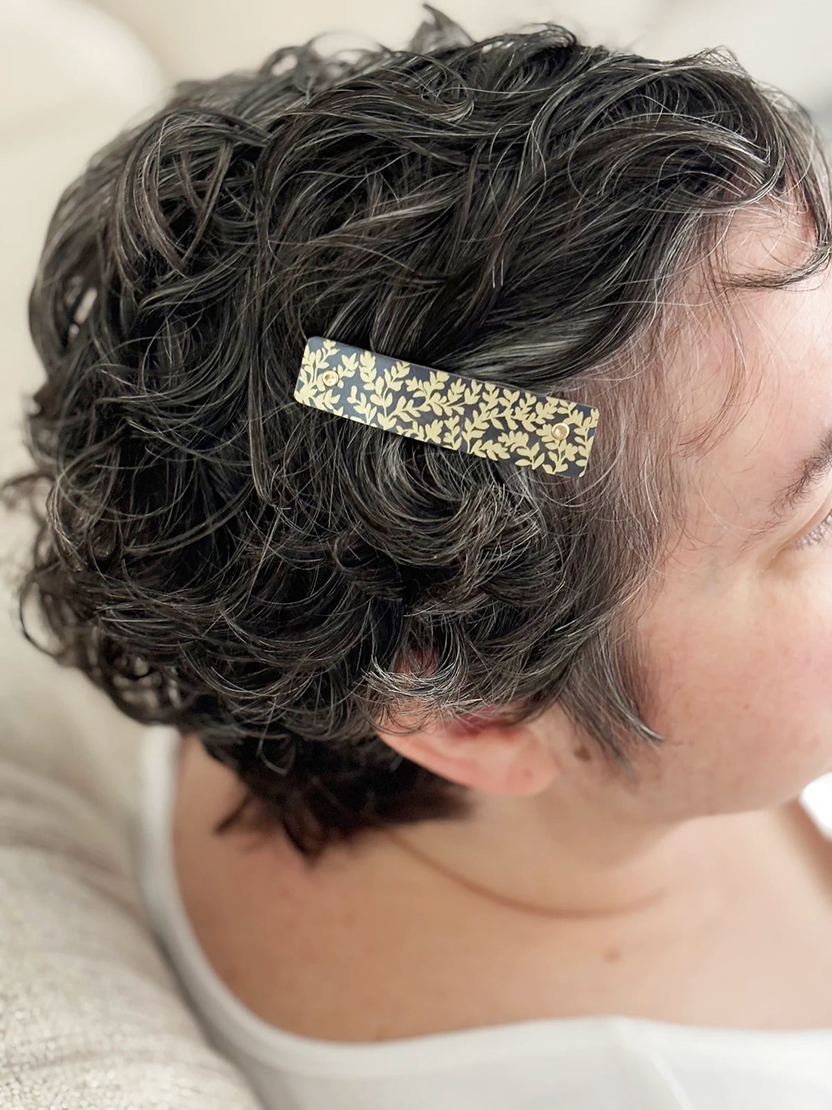 A close-up of a person's short, curly dark hair wearing a slim rectangular hair clip with a gold botanical pattern on a charcoal background.