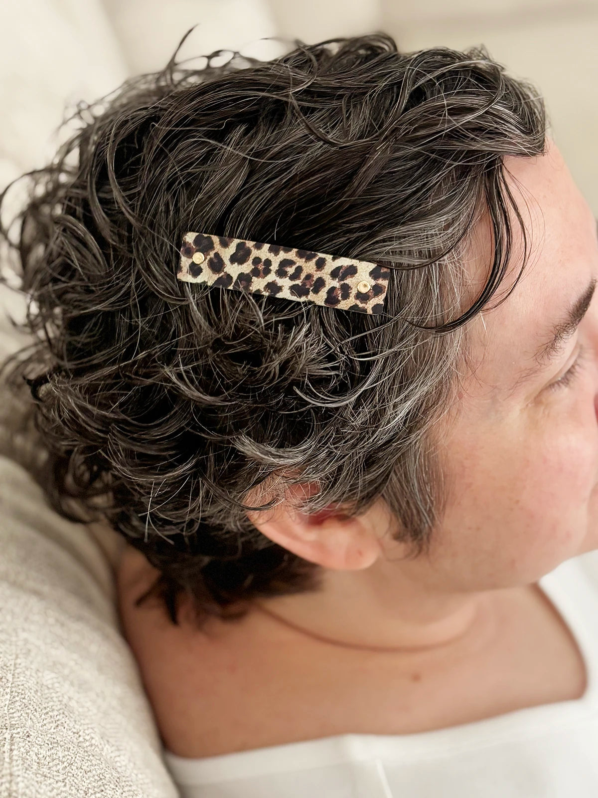 A close-up side view of a person with short, curly gray hair wearing a leopard print hair clip.
