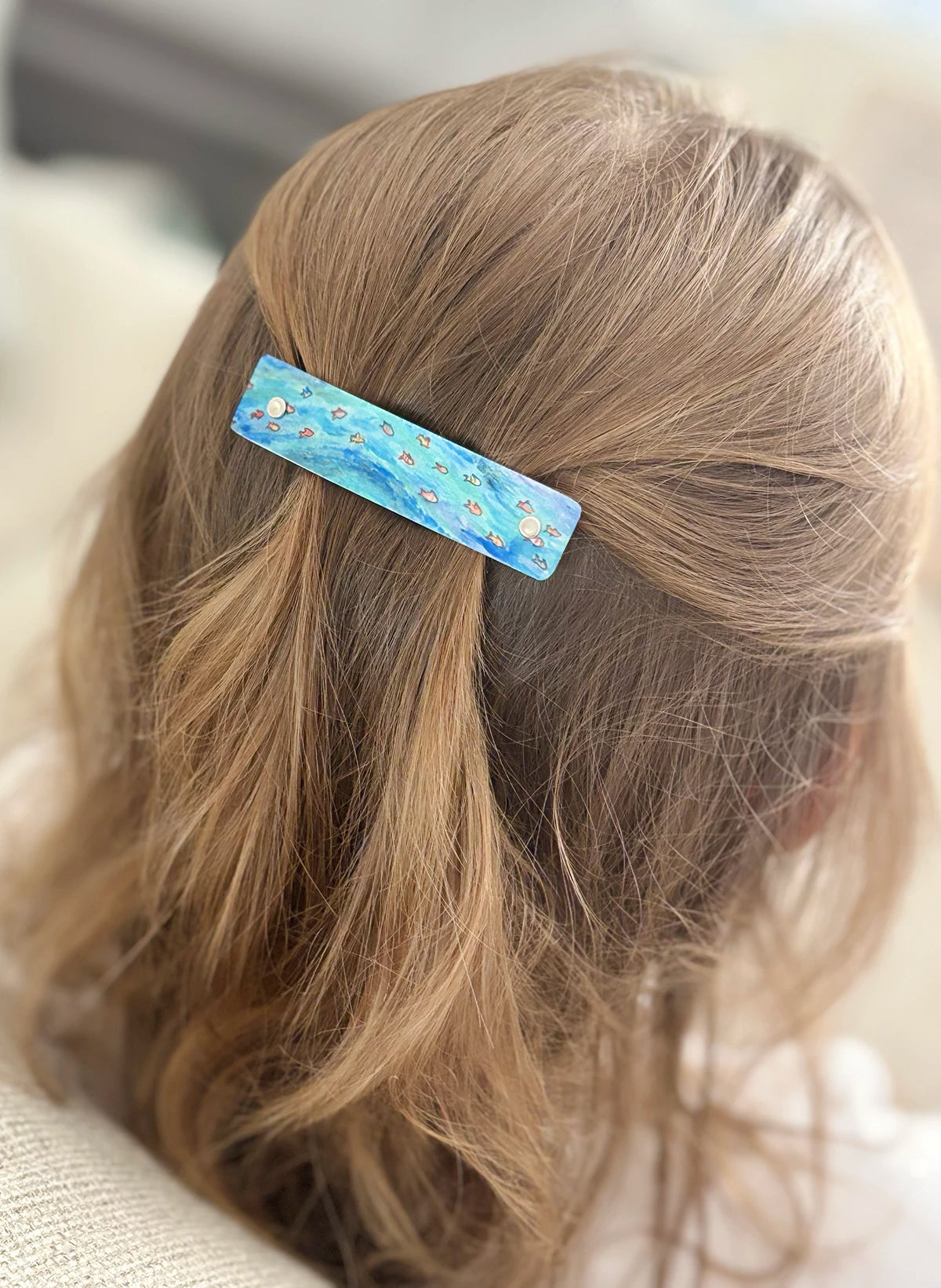 A close-up of a child's light brown hair secured with a small rectangular blue ocean hair clip decorated with tiny orange and yellow tropical fish.