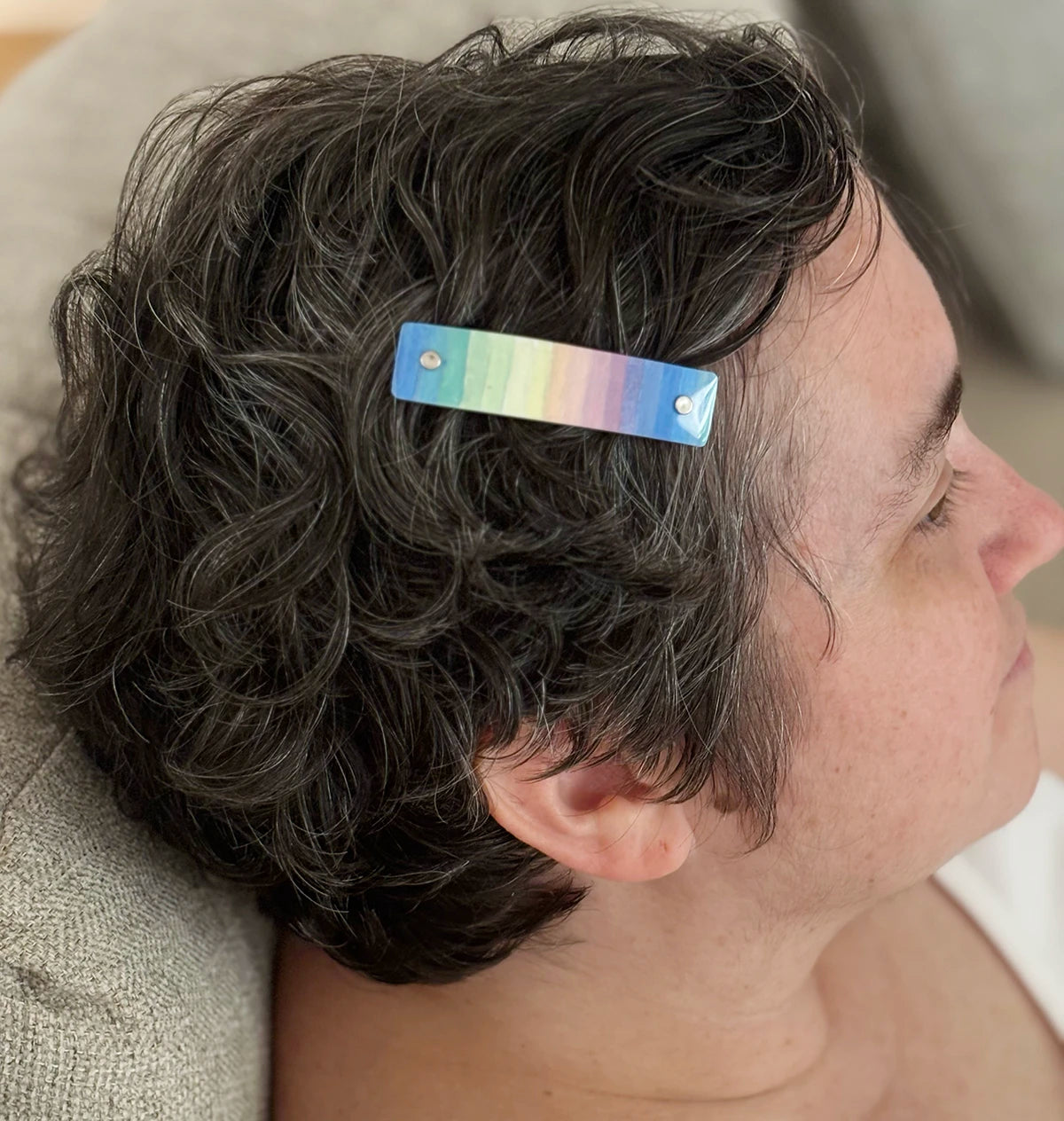 A close-up side profile of a person with short, wavy dark hair wearing a slim rectangular hair clip with a rainbow gradient finish.
