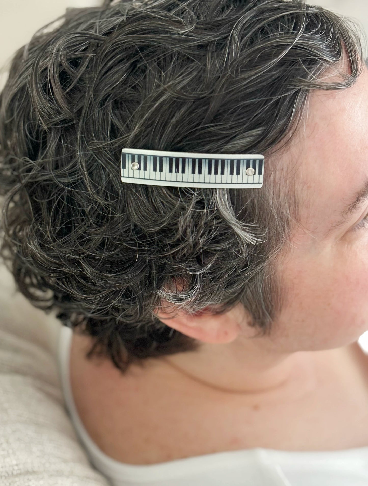 A close-up side profile of a person with short, curly gray hair wearing a silver hair clip with a piano-key design.