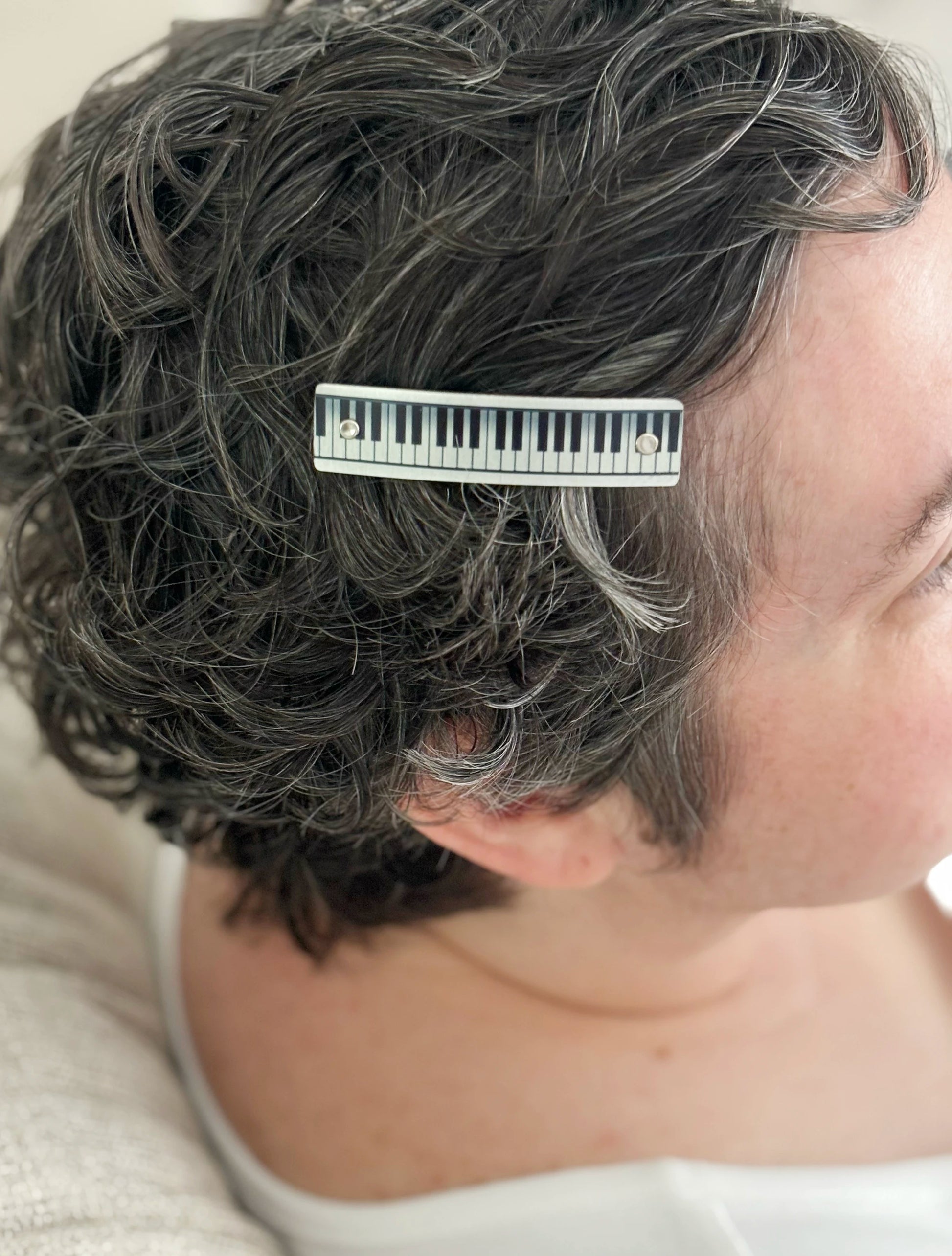 A close-up side profile of a person with short, curly gray hair wearing a silver hair clip with a piano-key design.