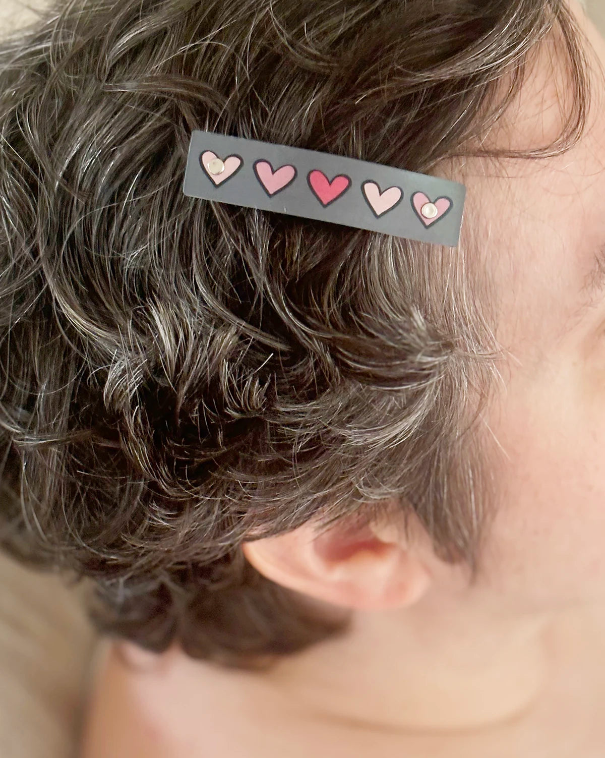 A close-up of a person's head showing short, curly gray-brown hair with a small rectangular hair clip holding pink hearts..