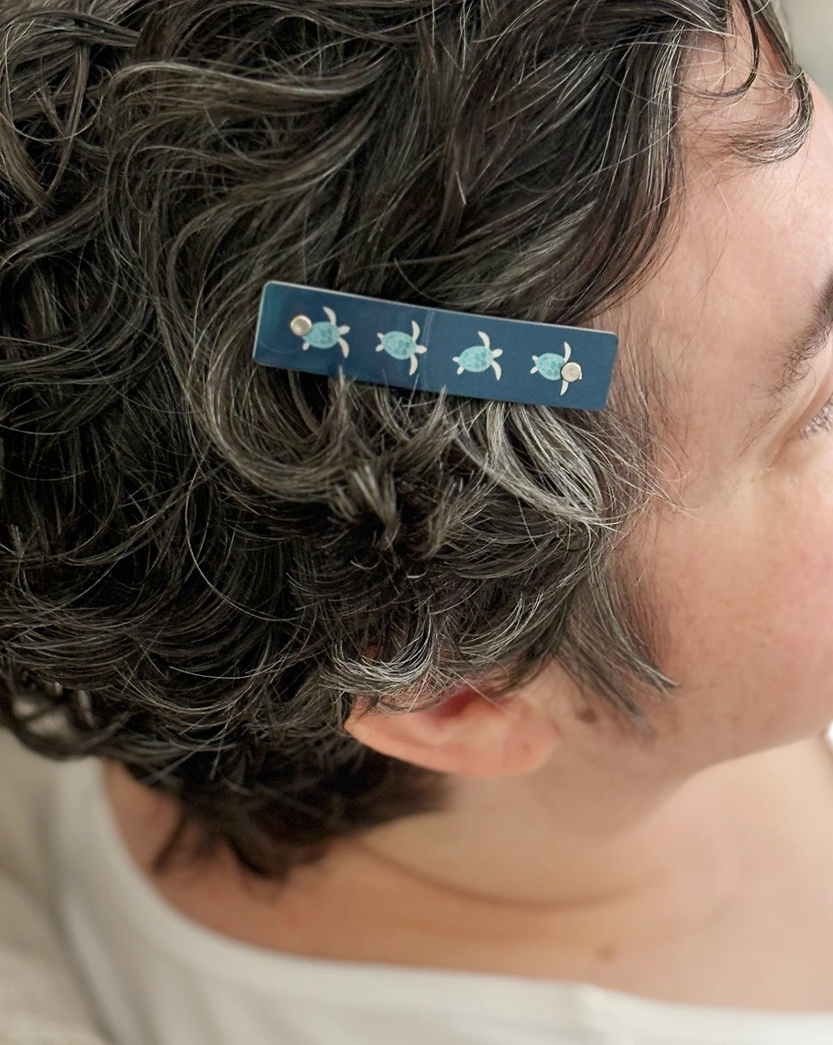 A close-up of a person's short, curly gray hair wearing a dark blue hair clip decorated with four small sea turtle motifs.