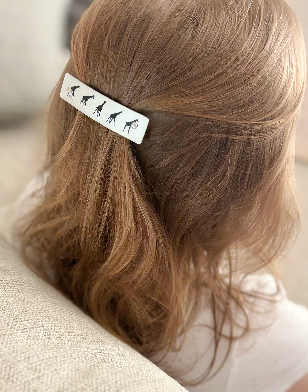 A close-up lifestyle shot of a person with light brown hair wearing a silver rectangular hair clip decorated with black silhouettes of giraffes.