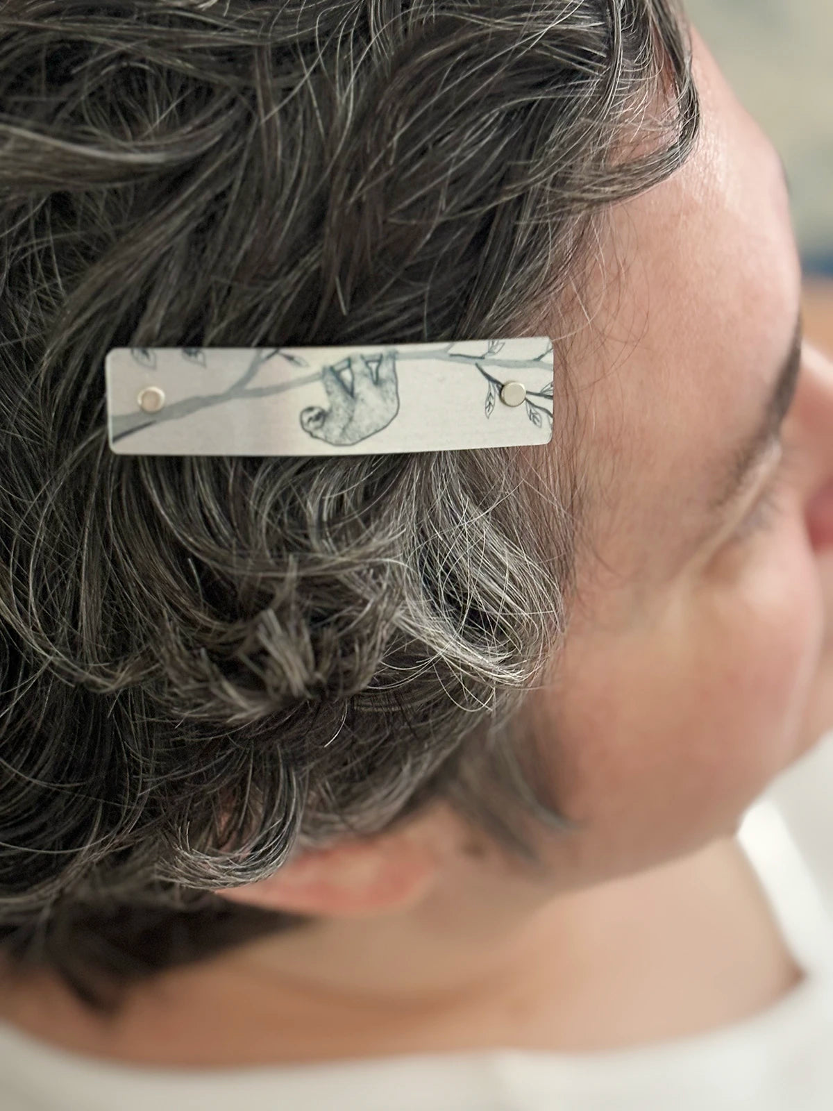 A close-up side view of a person with short, curly gray hair wearing a slim rectangular hair clip. The clip is silver with a sloth line drawing.