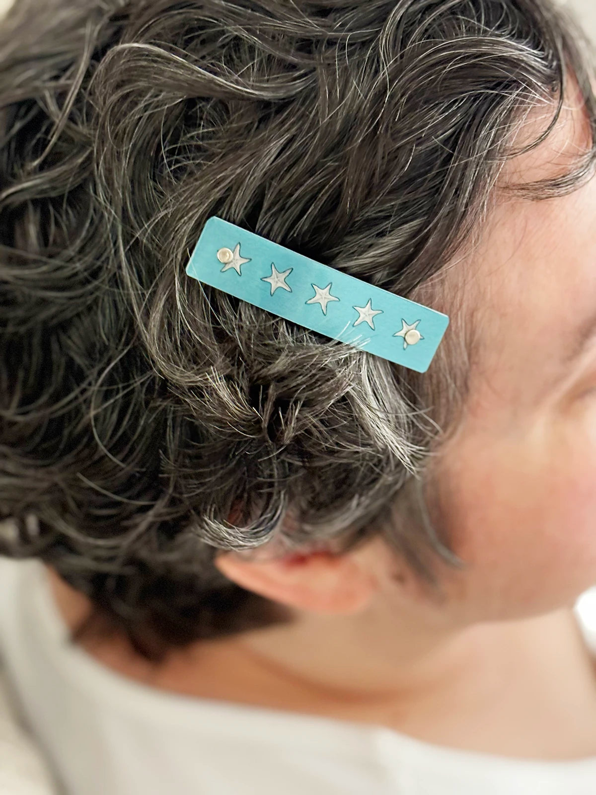 A close-up of a person's dark, wavy hair holding a light blue beach barrette hair clip decorated with small silver starfish.