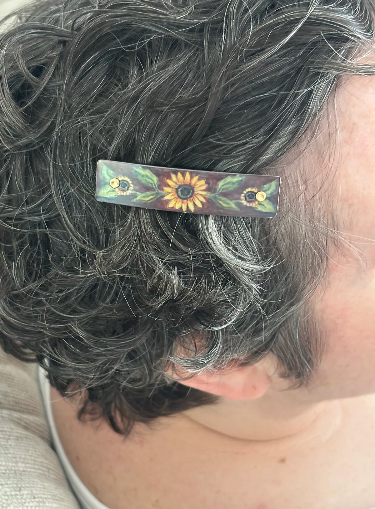 A close-up of a person's dark gray hair holding a rectangular hair clip decorated with painted sunflowers and green leaves.
