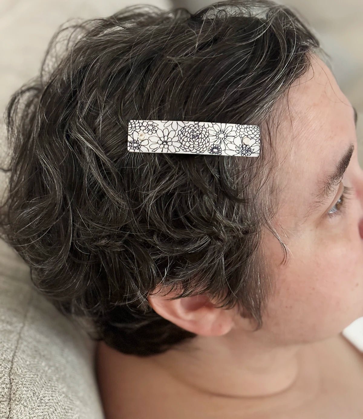 A close-up of a person's short, wavy dark hair with a small rectangular hair clip holding a section of hair back. The clip is silver with a black zinnia floral pattern.