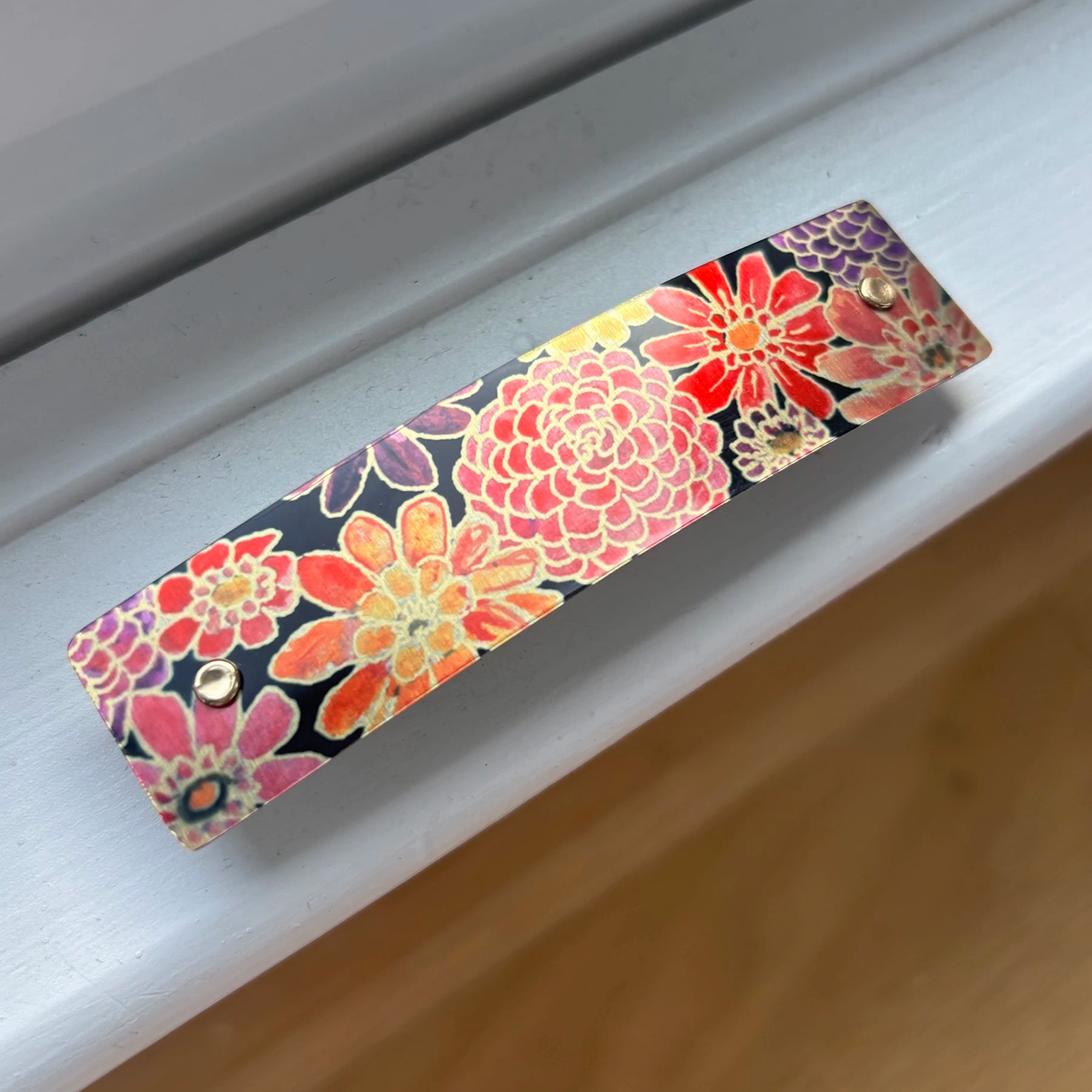 A rectangular handmade French barrette hair clip with colorful zinnia flowers on a black background, resting on a white window sill.