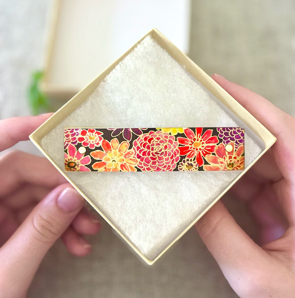 Red, orange, pink, yellow, and purple watercolor zinnias on a handmade French barrette in an open square box.