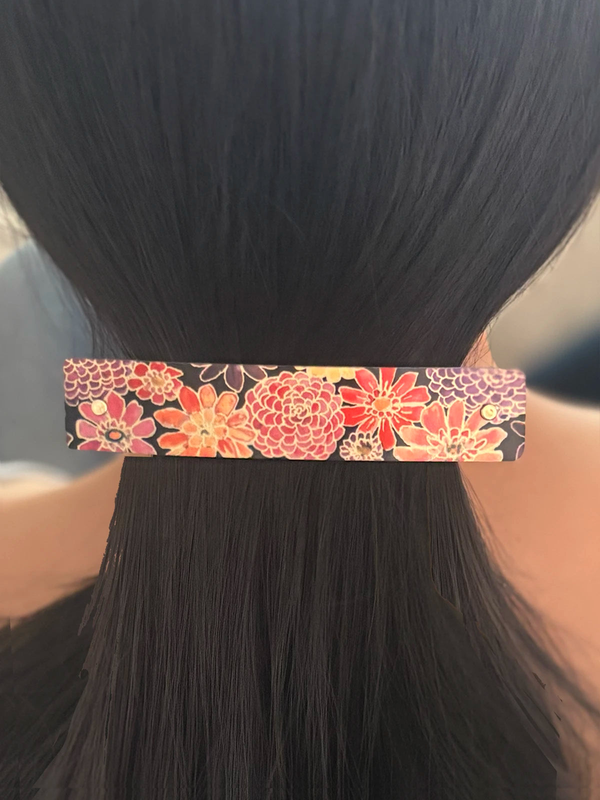 A close-up of a person's dark hair in a ponytail, showcasing a large rectangular hair clip with a colorful floral pattern in pink, orange, and gold on a black background.