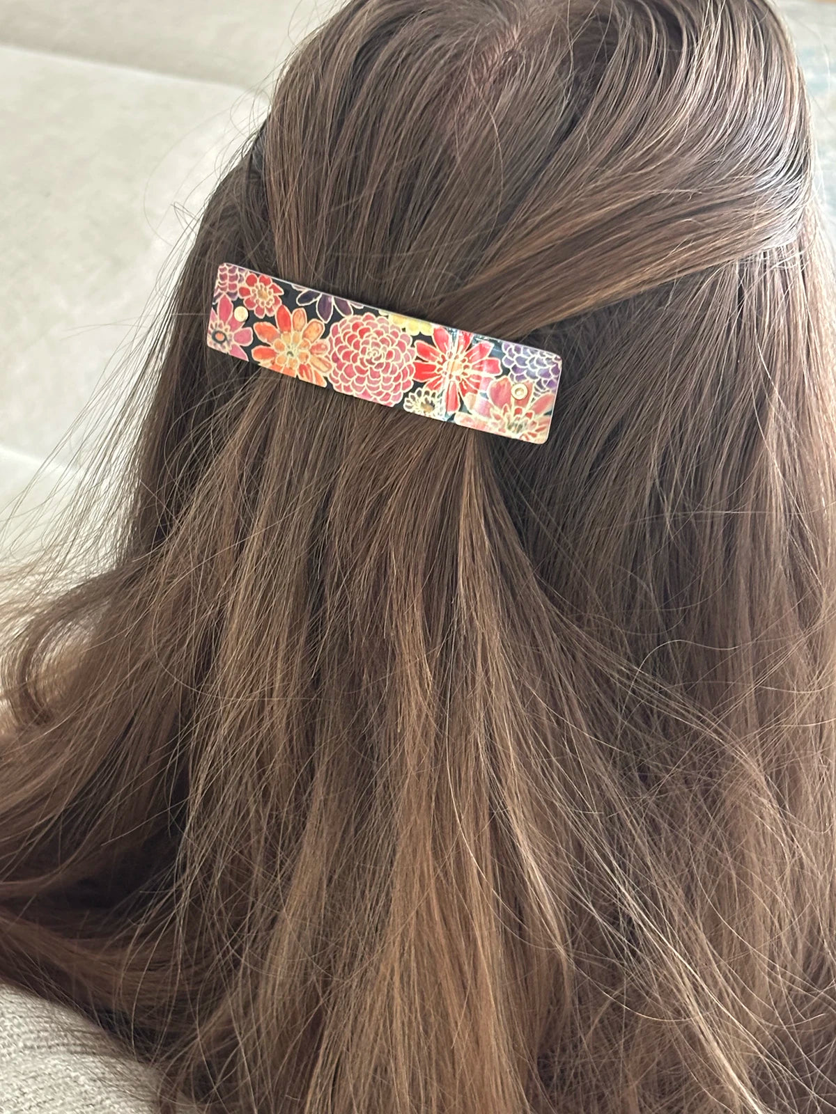 A close-up of a person's brown hair secured with a slim rectangular hair clip featuring a colorful floral pattern.