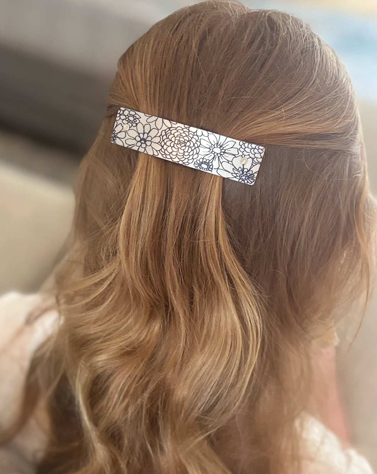 A close-up back view of a person with long, wavy blonde hair wearing a slim, rectangular hair clip with a black floral zinnia flower pattern on a silver background.