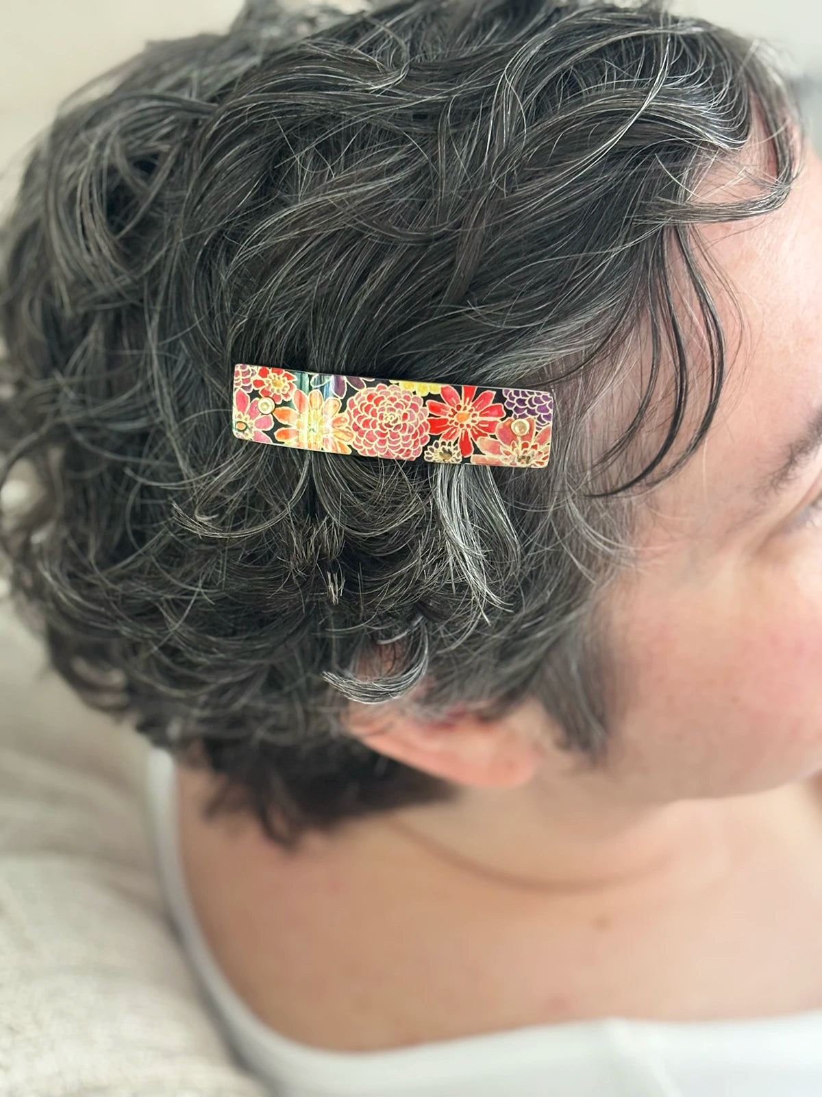 A close-up of a person with short, curly gray hair wearing a rectangular hair clip adorned with a colorful floral pattern.