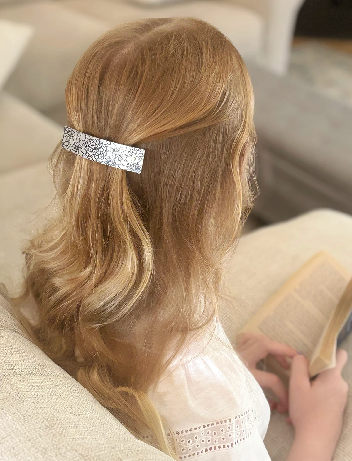 A blonde girl seen from behind wearing a silver hair clip with black floral drawn design, sitting on a beige couch and holding an open book.
