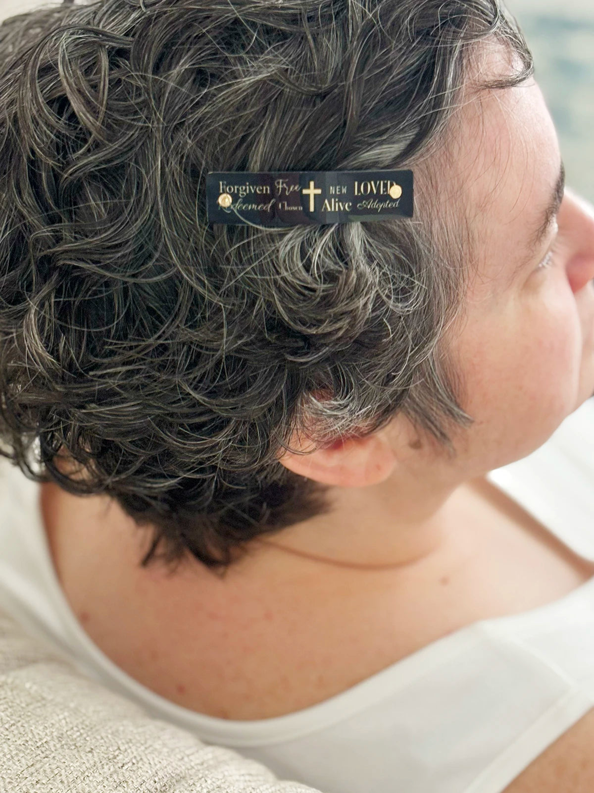 A close-up side profile of a person with short, curly gray hair wearing a small black hair clip. The clip has gold lettering that reads 'Forgiven Free Redeemed Chose New Alive Adopted' and a gold cross in the center.
