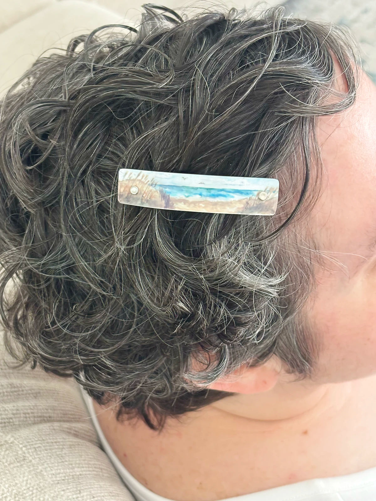 A close-up of a person's short, curly gray hair with an extra small handmade hair clip holding it back. The clip has a light blue and white beach scene in watercolor impressionist style.