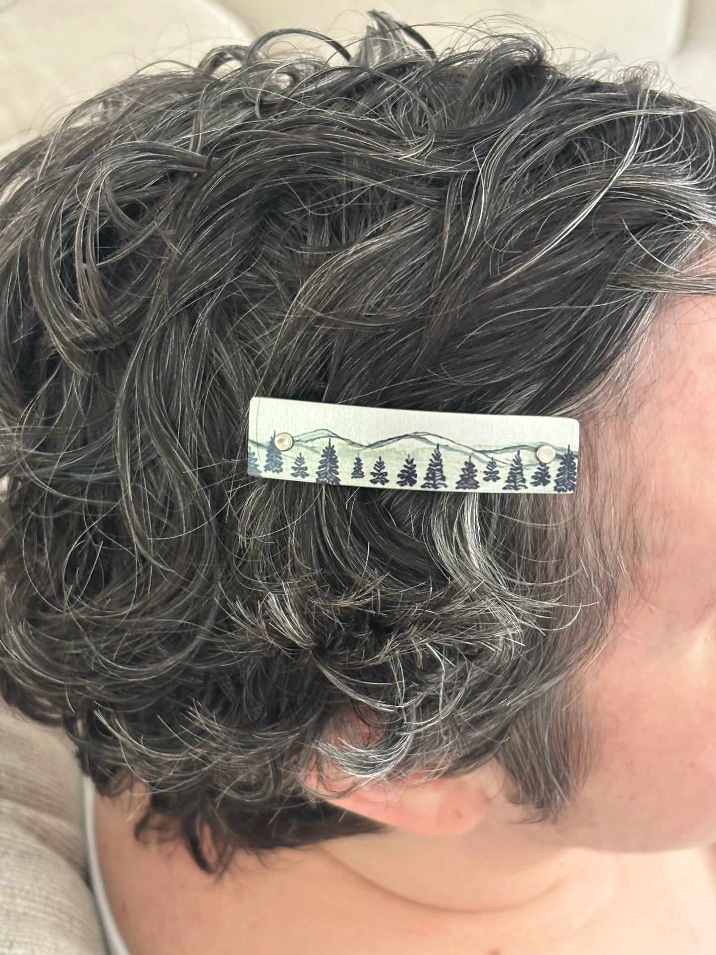 A close-up of a person's short, curly gray hair wearing a silvery hair clip decorated with a black forest and mountain pattern.