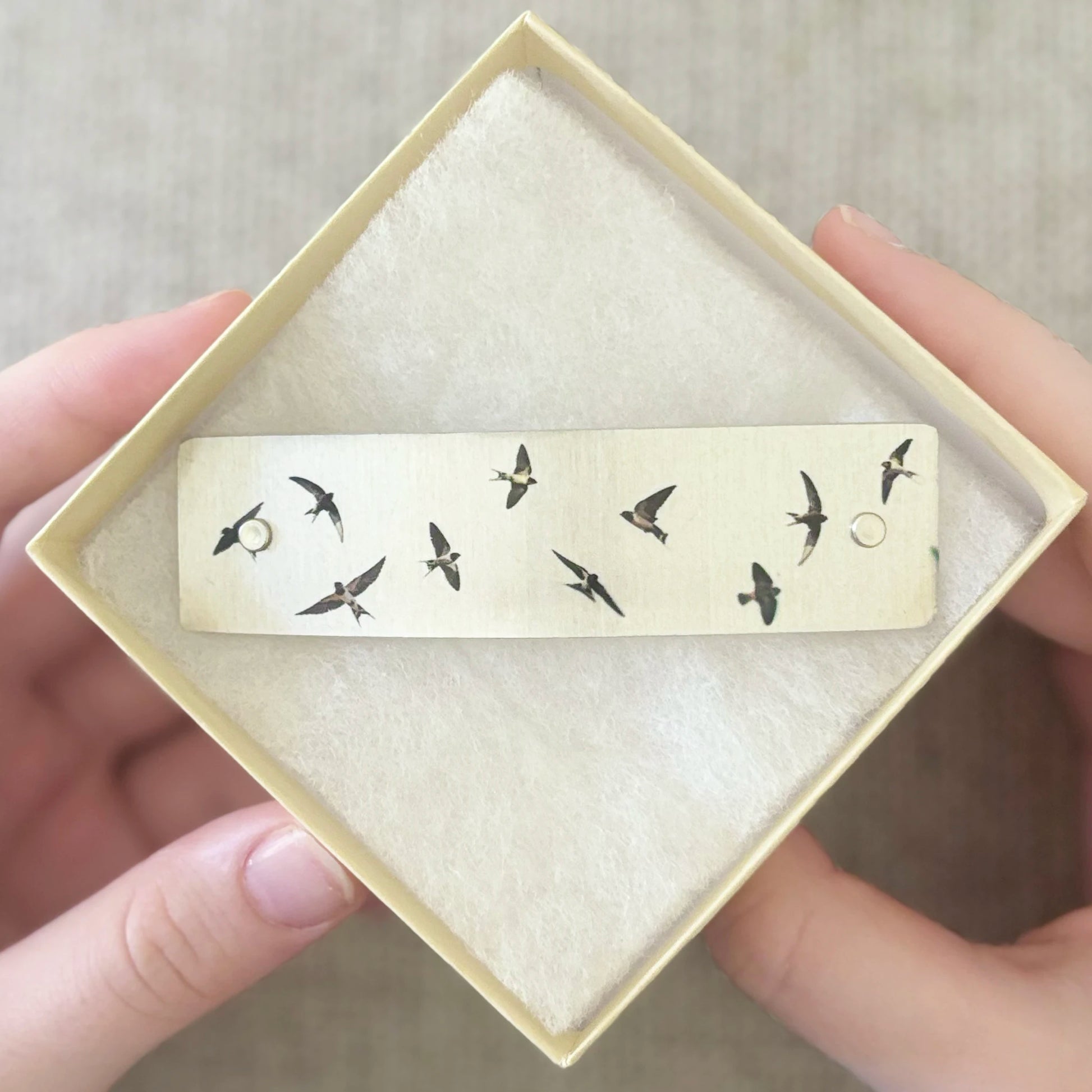A rectangular cream-colored barrette with black bird silhouettes, held in a square cream gift box with a soft insert. A person's hands are visible holding the box against a neutral fabric background.