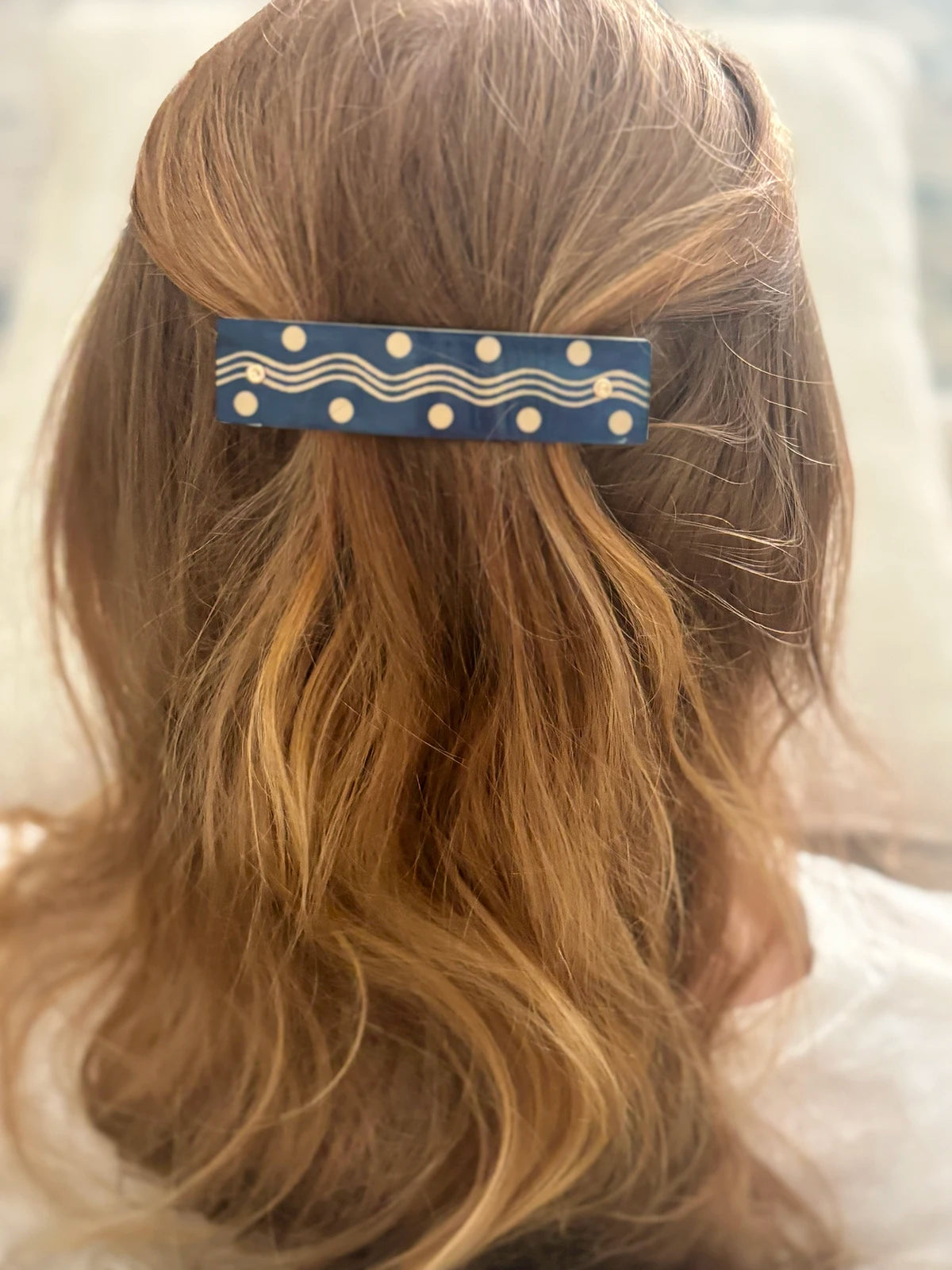 A close-up back view of a person with long, wavy strawberry blond hair wearing a navy blue hair clip decorated with gold-colored wavy lines and polka dots.