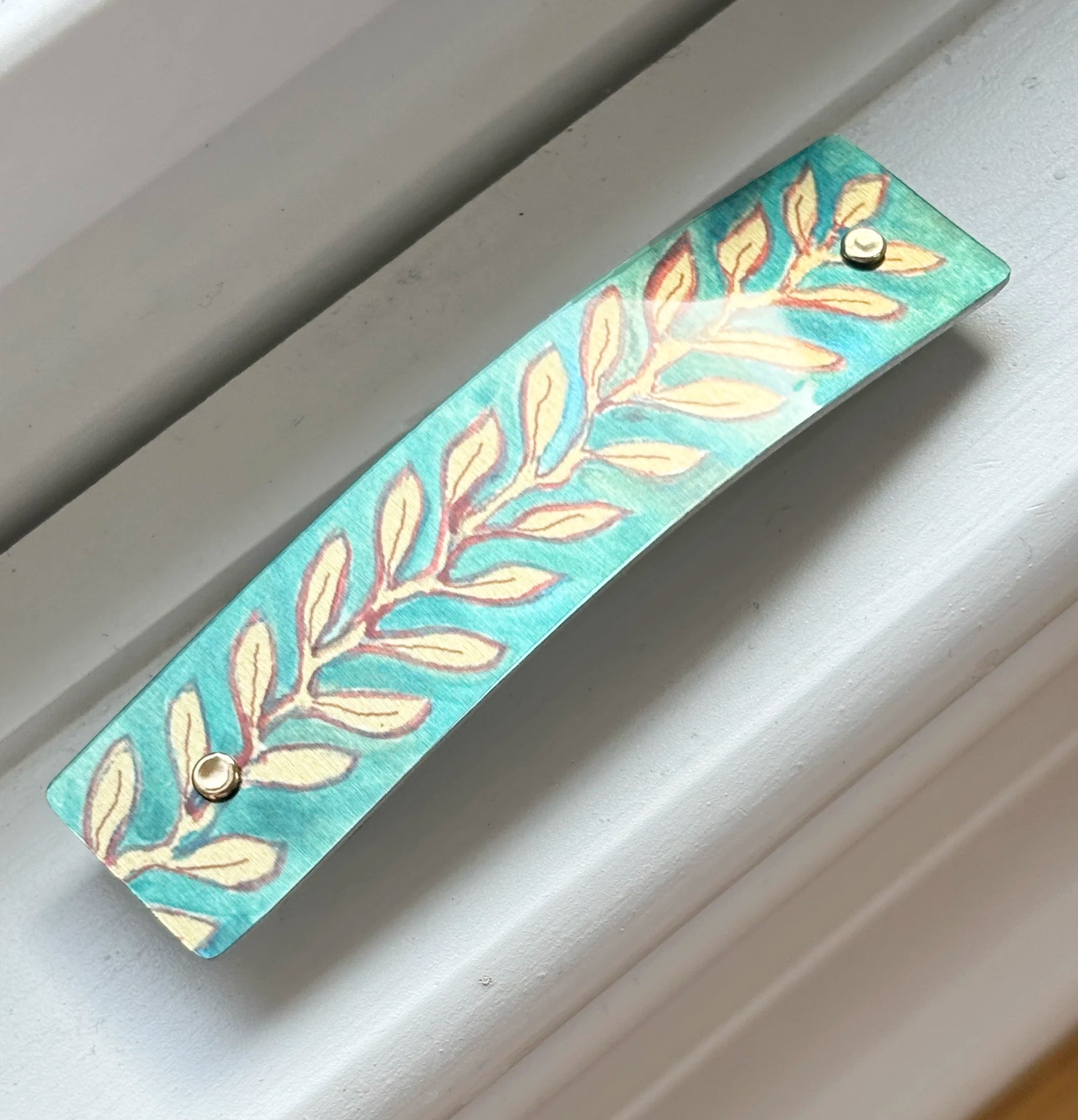 A rectangular turquoise metal barrette with a hand-painted leaf pattern in gold and brown, secured with two small gold rivets, shown resting on a white window sill.