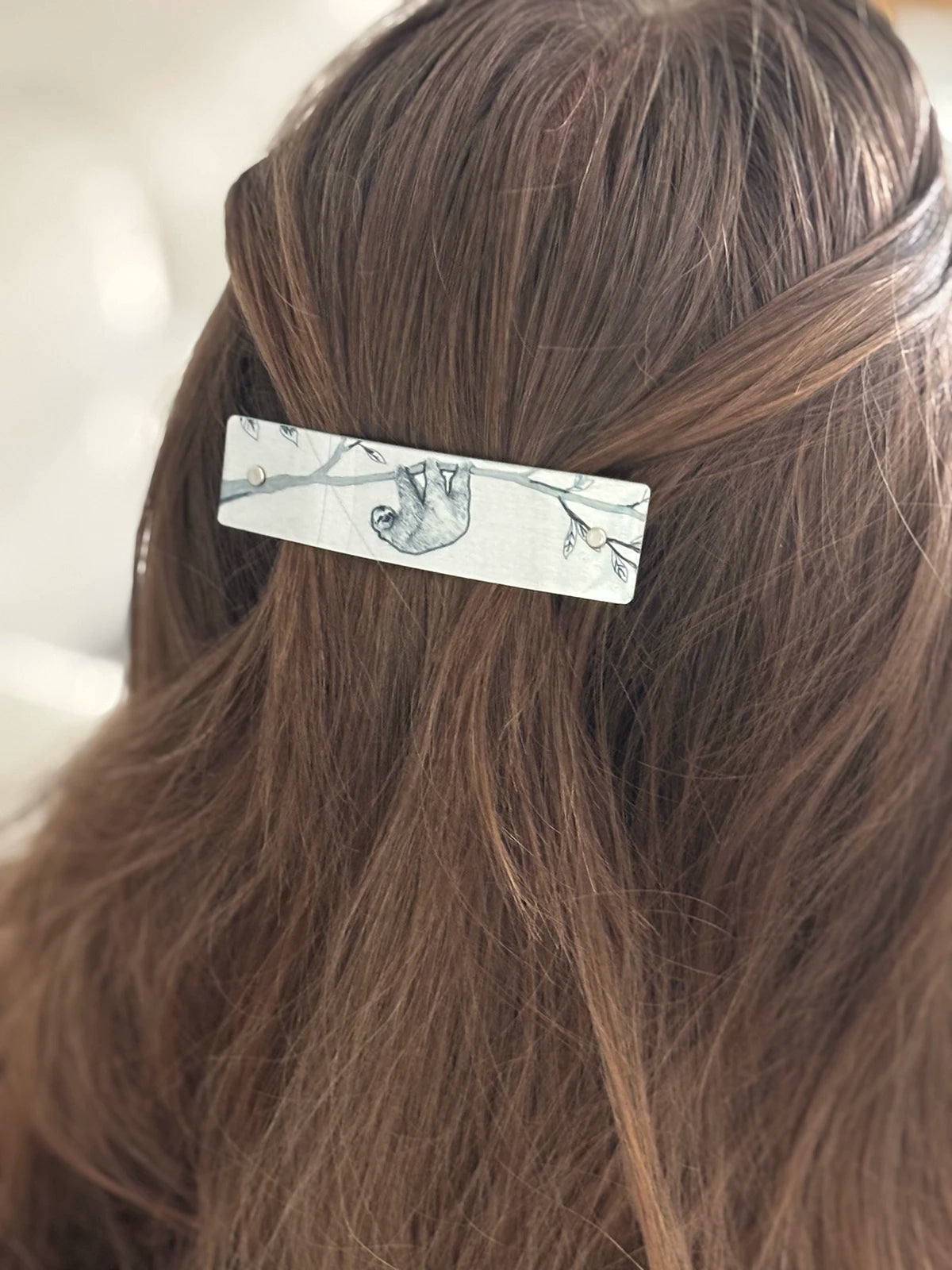 A close-up of a person's brown hair secured with a slim rectangular hair clip featuring a silver background and a black line drawing of a sloth.