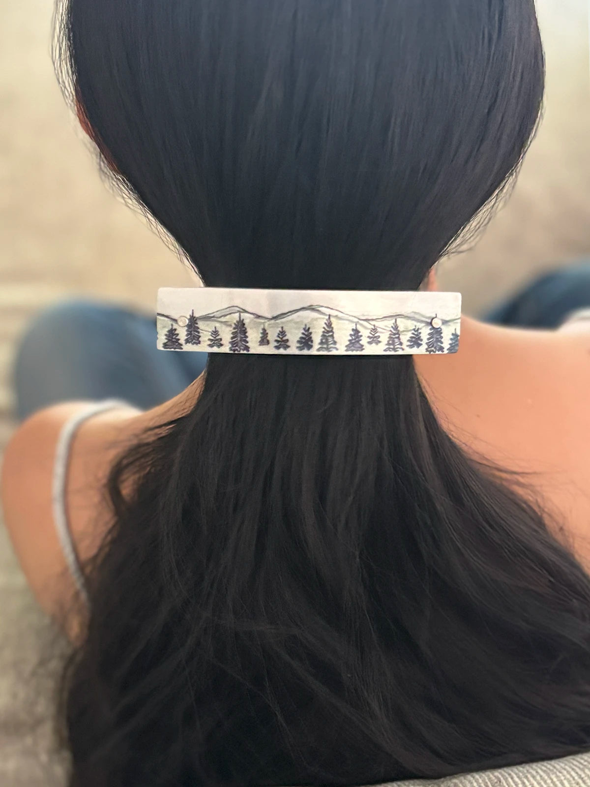 A close-up back view of a person with long black hair secured in a low ponytail, with a rectangular silver hair clip decorated with an artistic black mountain and pine tree pattern.