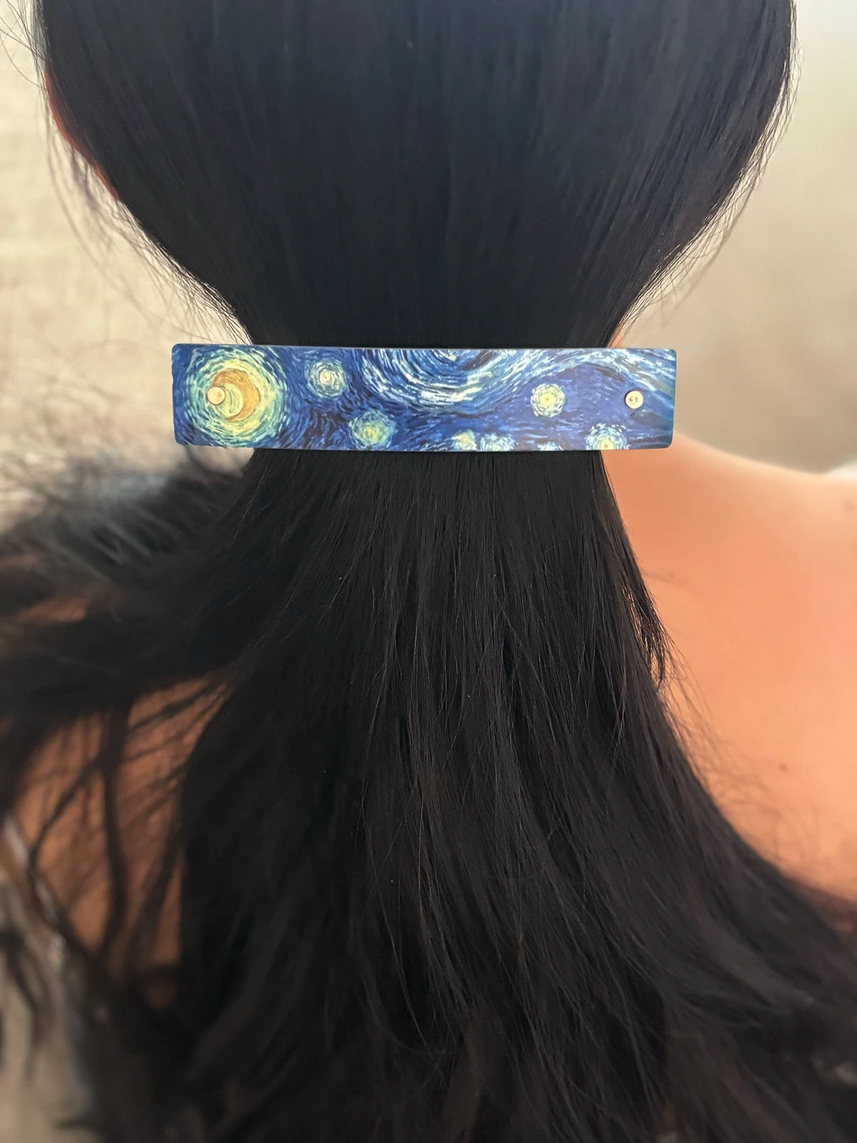A close-up of a person's dark hair tied back in a low ponytail, secured with a rectangular barrette featuring a blue and yellow Starry Night painting design.