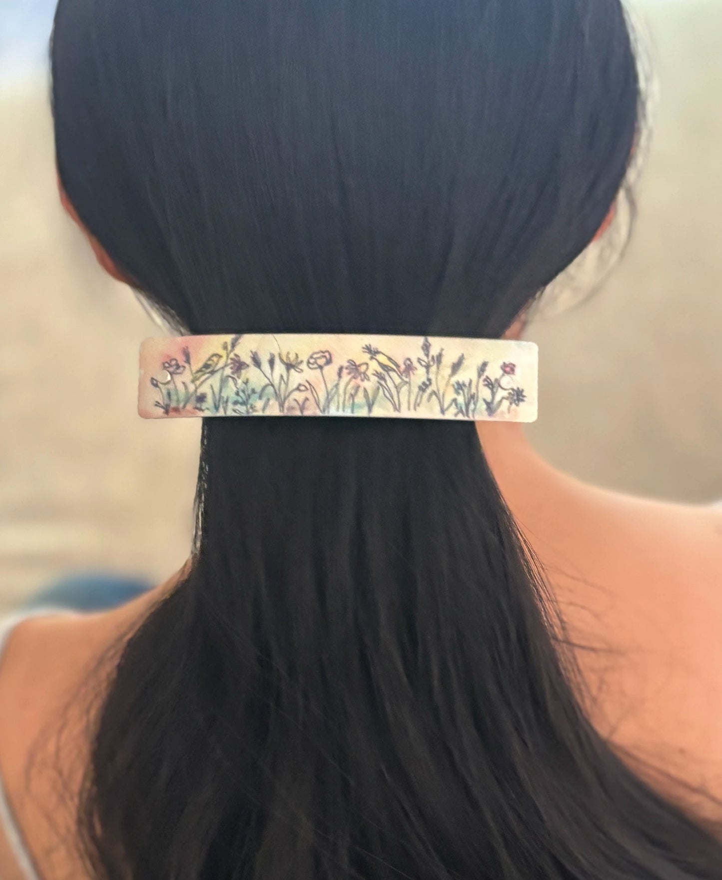 A close-up back view of a person with long black hair wearing a slim handmade hair clip decorated with a colorful wildflower watercolor.