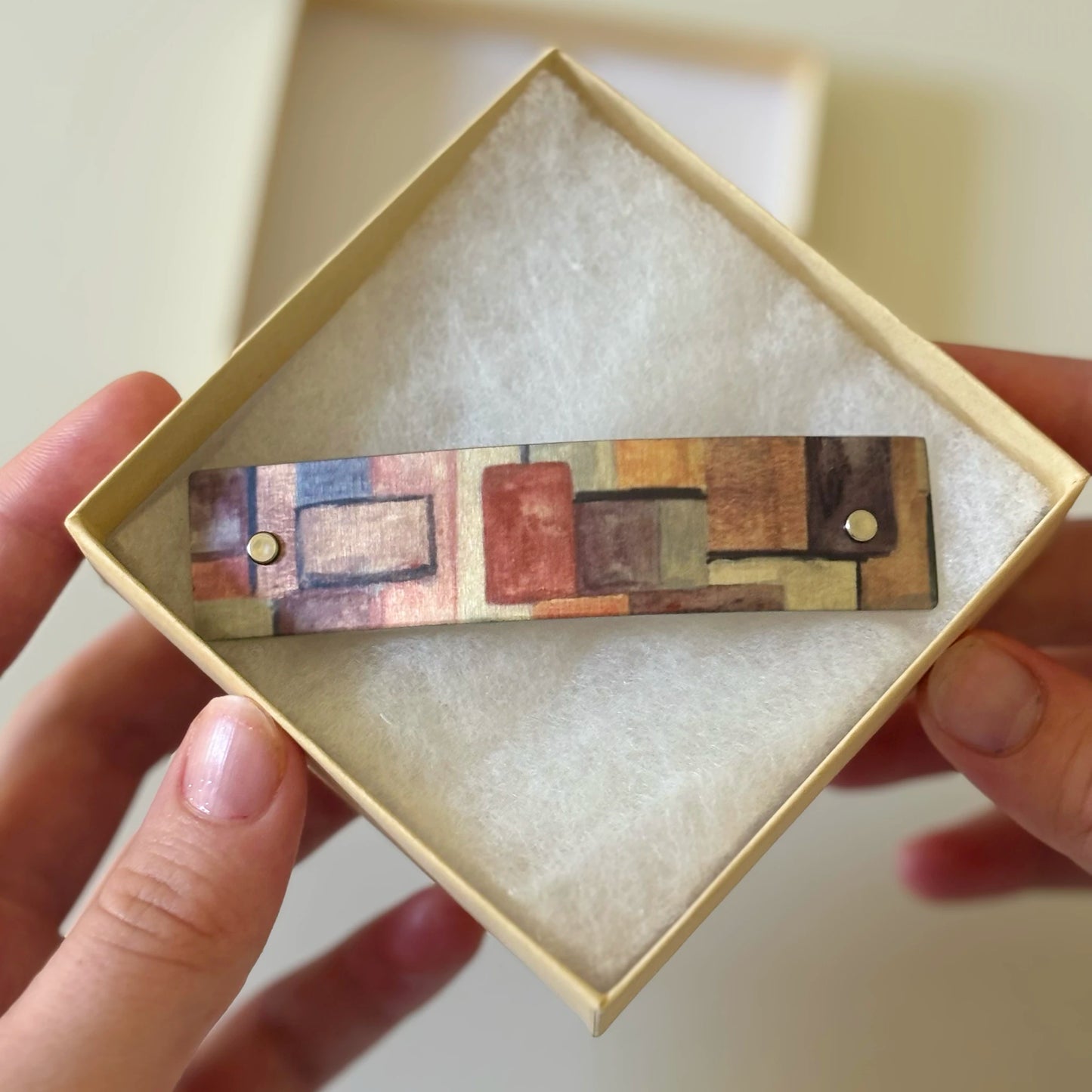 A rectangular aluminum French barrette with an  original abstract geometric pattern in earthy tones, held in a person's hands inside a light beige presentation box with white lining.