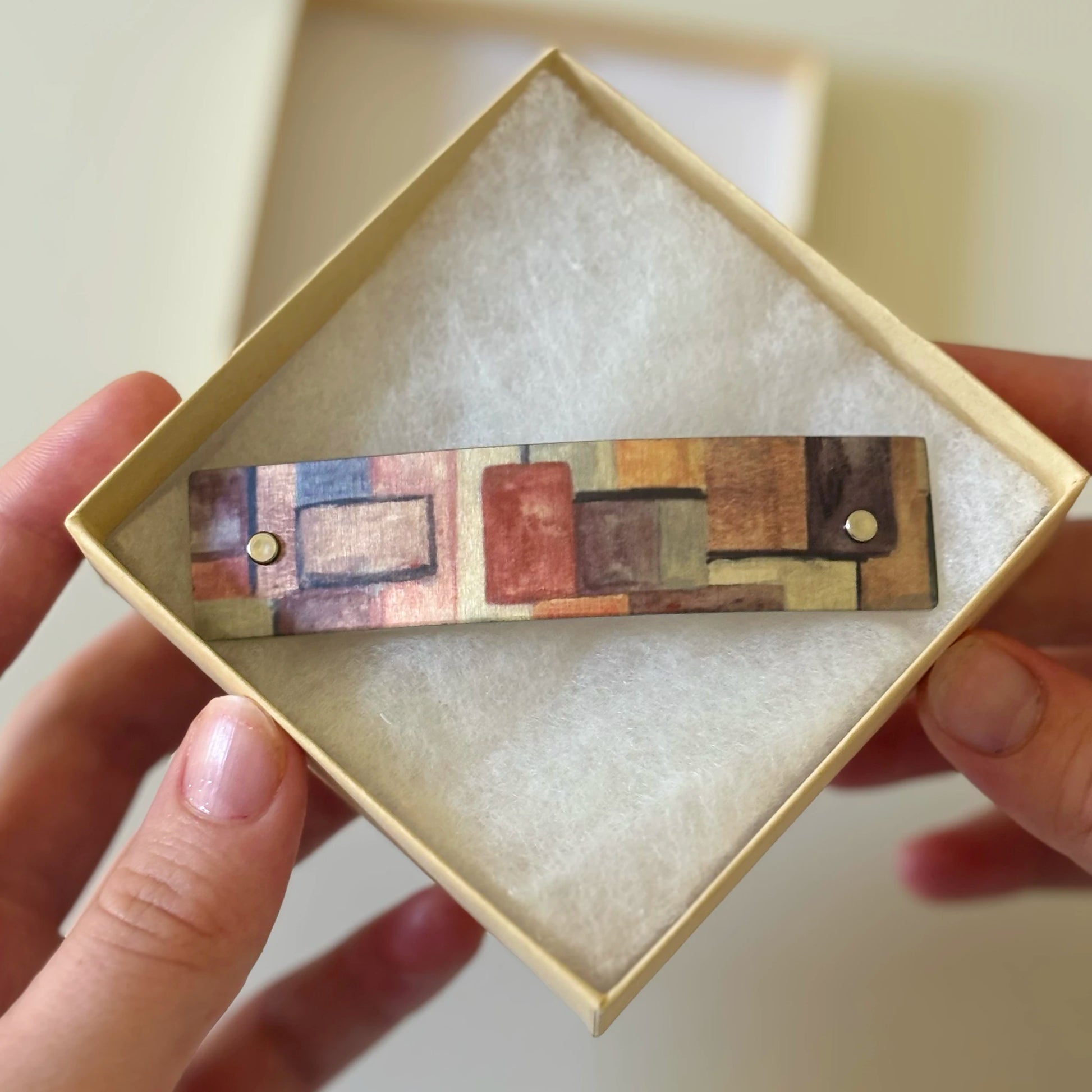 A rectangular aluminum French barrette with an  original abstract geometric pattern in earthy tones, held in a person's hands inside a light beige presentation box with white lining.