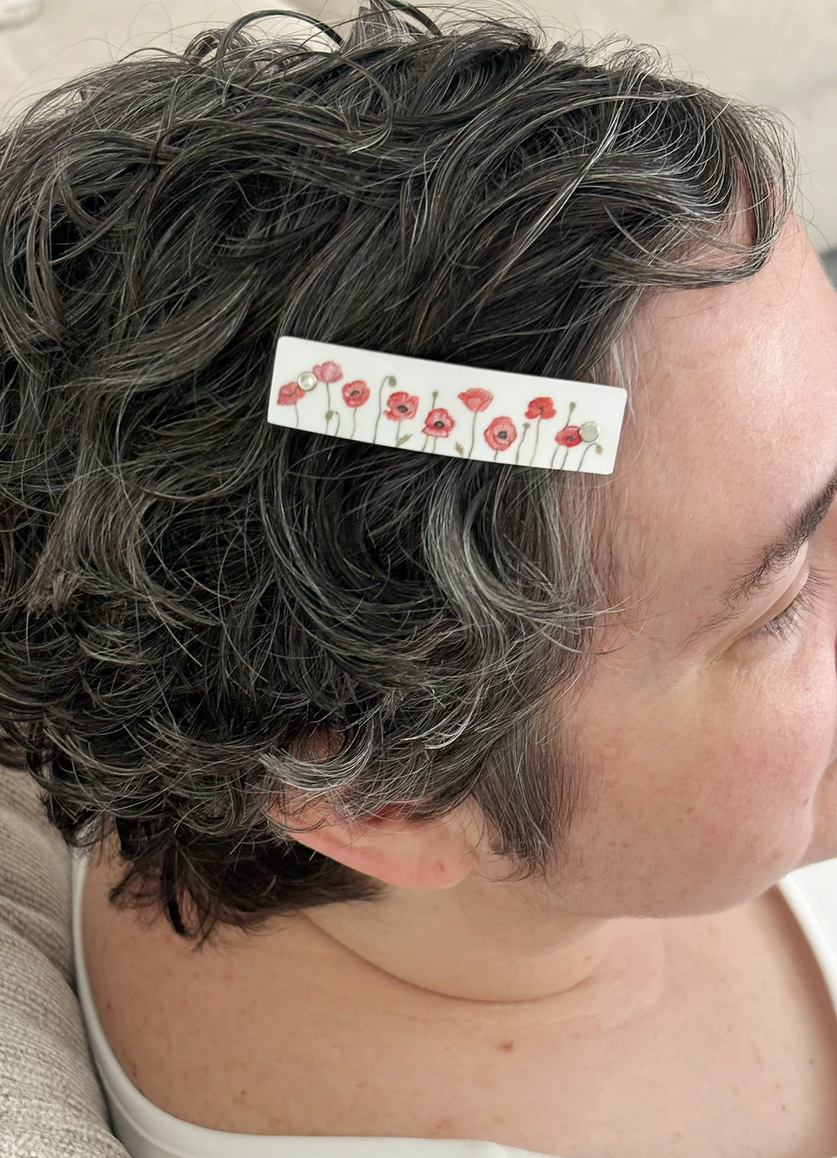 A close-up side profile of a person with short, curly gray hair wearing a white hair clip decorated with a red poppy floral watercolor design by Robbin V. Davis.