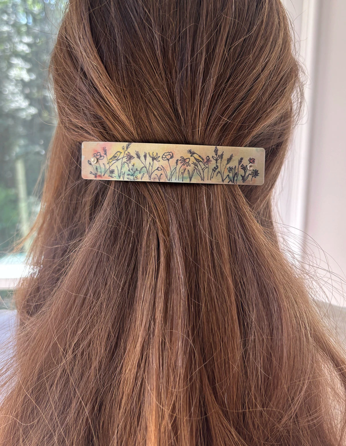A close-up lifestyle shot of a woman's brown hair secured with a rectangular boutique hair clip decorated with an original watercolor wildflower and bird pattern, set against a softly blurred indoor background.