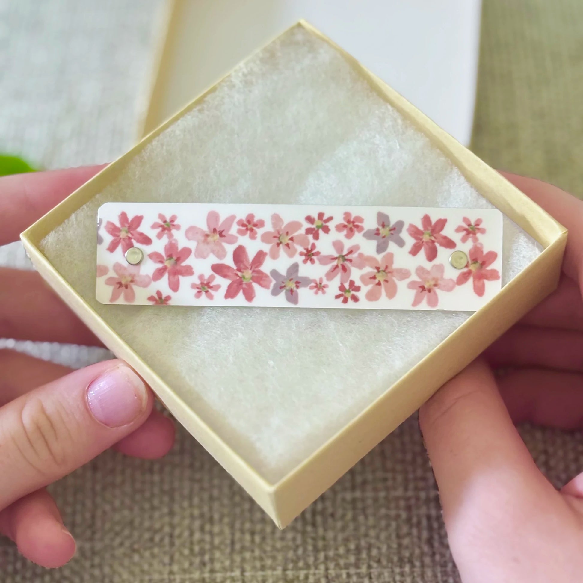 A close-up of hands holding a small cream-colored square gift box with a floral hair clip inside inside. The hair clip features a pattern of pink blossoms with a white background.